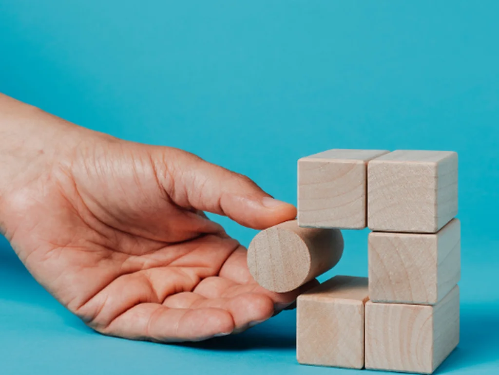 Titel: Strategische Problemlösung in der IT-Beratung

Beschreibung: Eine Hand platziert einen runden Holzblock in einen Würfel aus Holzklötzen, der vor einem hellblauen Hintergrund steht. Dieses Bild symbolisiert innovative Problemlösungen in einer IT-Age