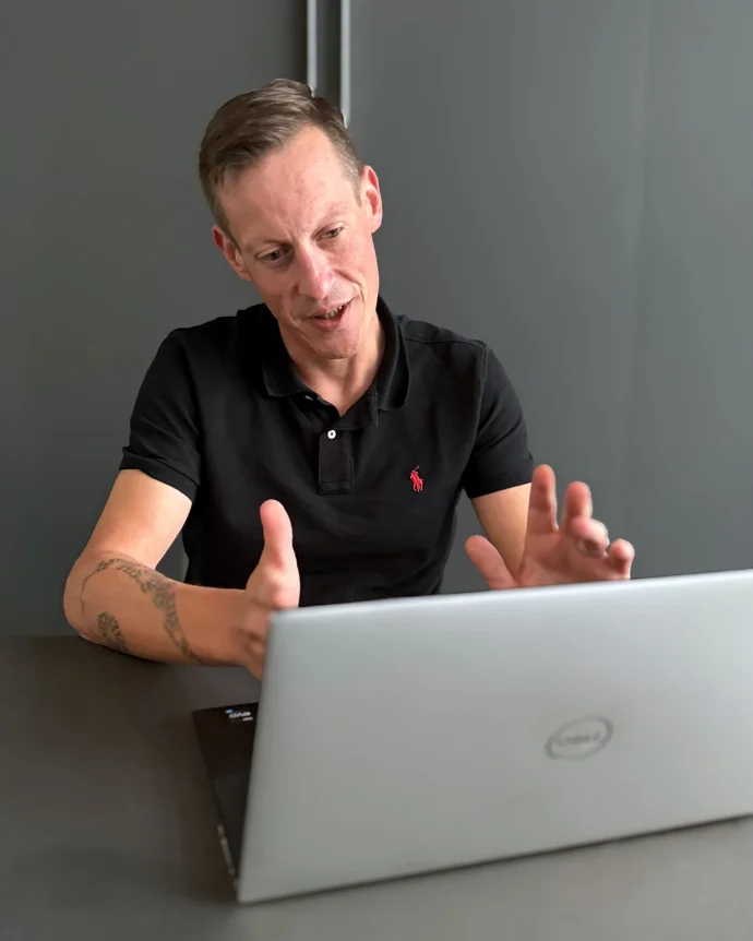 Person in schwarzem Polohemd gestikuliert vor einem Laptop im Gespräch.