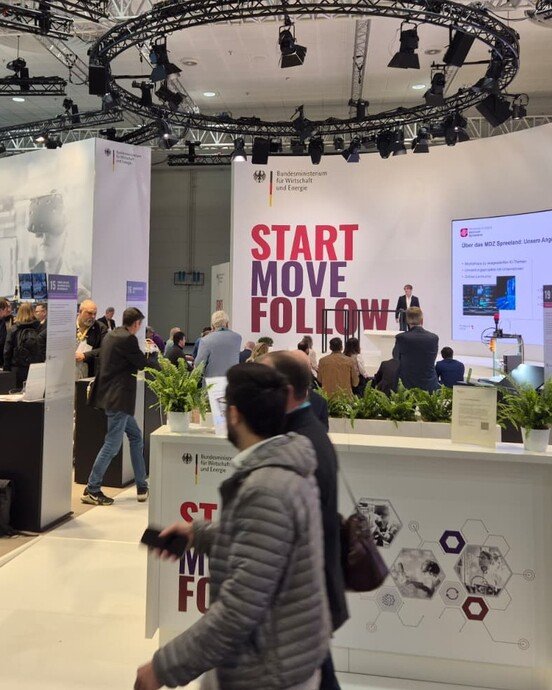 Messestand mit Besuchern und Präsentationen, beleuchtet von kreisförmiger Deckenbeleuchtung. Aufschrift: "START MOVE FOLLOW".