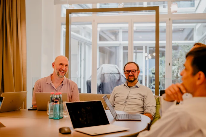 **Bildbeschreibung 1:** In einem modernen Büro einer IT-Agentur sitzen mehrere Mitarbeiter rund um einen Konferenztisch und nutzen ihre Laptops. Sie scheinen in ein kreatives Brainstorming oder eine Teambesprechung vertieft zu sein. Die helle und einladen