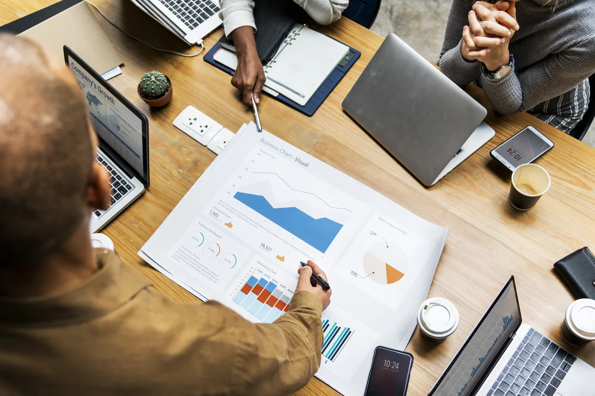 **Bildbeschreibung: Strategiebesprechung in IT-Agentur**

Ein dynamisches Team von Fachleuten in einer IT-Agentur führt eine Strategiebesprechung durch. Auf dem Holztisch sind verschiedene moderne Geräte wie Laptops und Tablets zu sehen, zusammen mit deta