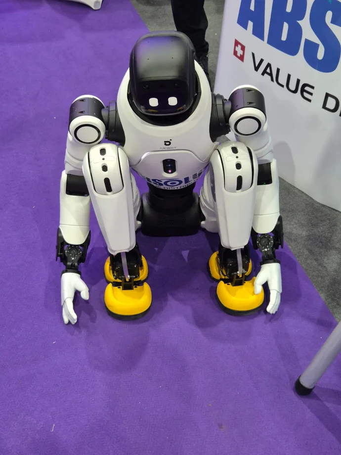 Ein humanoider Roboter kniet auf einem lila Teppich, mit weißen Armen und gelben Füßen.