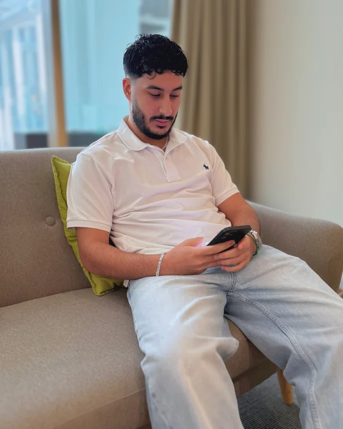Ein Mann sitzt auf einem Sofa und schaut auf sein Smartphone. Er trägt ein weißes Poloshirt und Jeans.