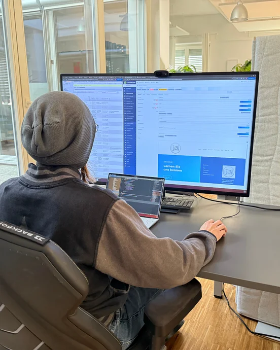 1. **Mitarbeiter einer IT-Agentur bei der Arbeit:** Ein konzentrierter IT-Experte arbeitet an einem modernen Arbeitsplatz mit zwei Bildschirmen. Der größere Monitor zeigt Unternehmenssoftware, während der Laptop für Programmieraufgaben genutzt wird. Diese