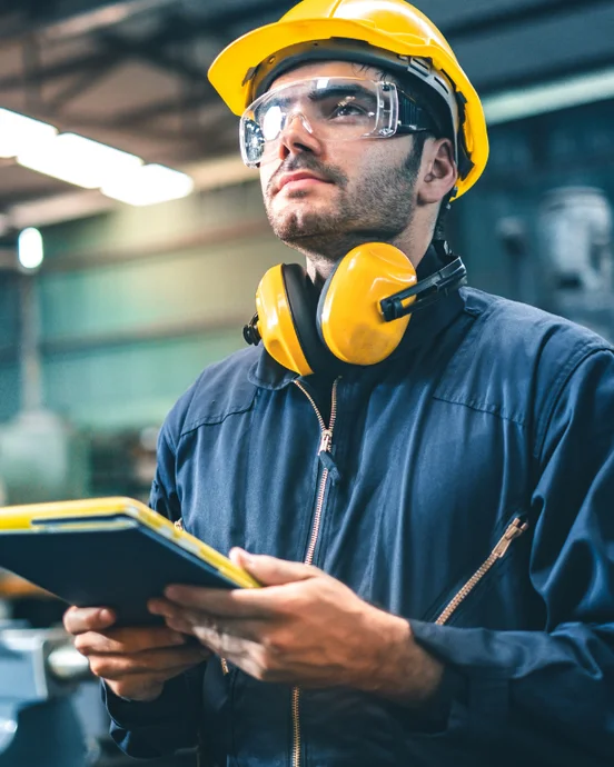 Ingenieur mit Sicherheitsausrüstung inspiziert Fertigungsanlage, hält ein Tablet, trägt Helm und Gehörschutz, industrielles Umfeld.
