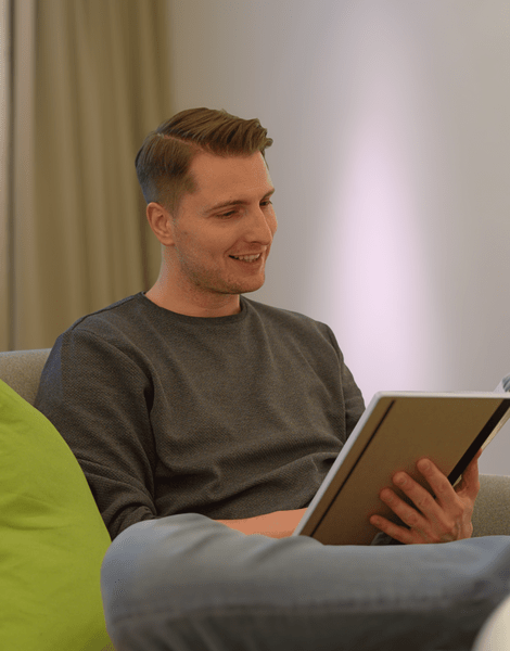 Eine Person sitzt auf einem Sofa, liest in einem Buch und lächelt, ein grünes Kissen ist neben ihr.