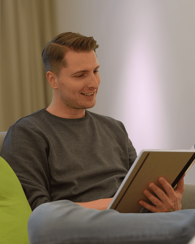 Eine Person sitzt auf einem Sofa, liest in einem Buch und lächelt, ein grünes Kissen ist neben ihr.