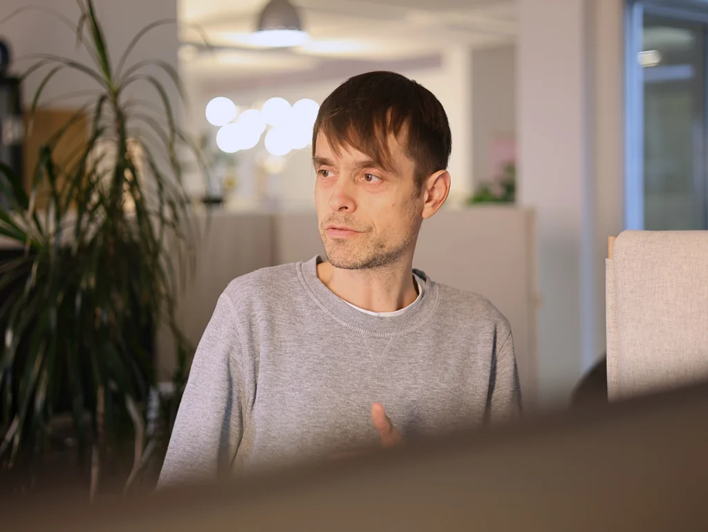 Eine Person in Freizeitkleidung sitzt in einem modernen Büro, im Hintergrund ist eine Pflanze und verschwommenes Licht zu sehen.