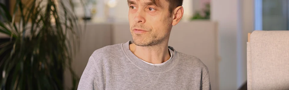 Eine Person in Freizeitkleidung sitzt in einem modernen Büro, im Hintergrund ist eine Pflanze und verschwommenes Licht zu sehen.