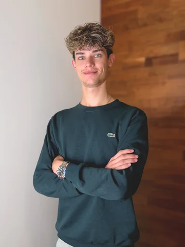 Eine Person in weißem Hemd steht vor einer hölzernen Wand und blickt in die Kamera.