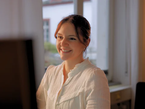 Eine Frau sitzt lächelnd vor einem Computer in einem hellen Raum mit Fenstern.