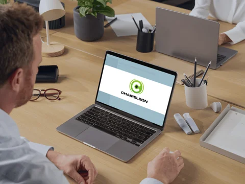 Mann arbeitet an Laptop mit Chameleon-Logo auf dem Bildschirm, im modernen Büroambiente mit Schreibtisch und Pflanzen.