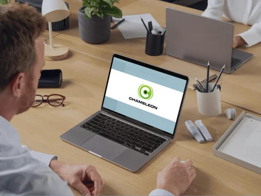 Mann arbeitet an Laptop mit Chameleon-Logo auf dem Bildschirm, im modernen Büroambiente mit Schreibtisch und Pflanzen.