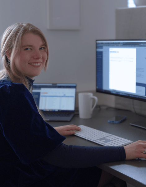 Person am Computertisch arbeitet an zwei Monitoren; lächelt zur Kamera. Moderne Büroumgebung.