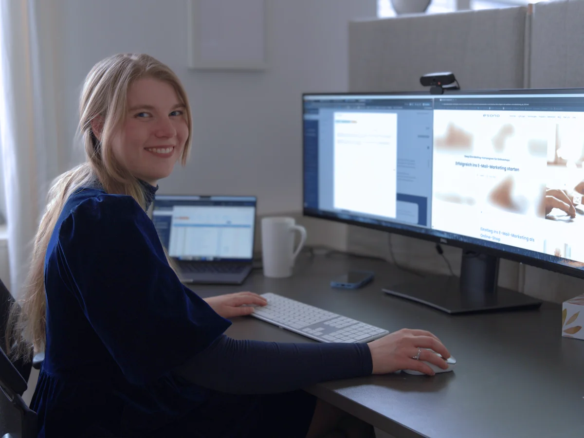 Person am Computertisch arbeitet an zwei Monitoren; lächelt zur Kamera. Moderne Büroumgebung.
