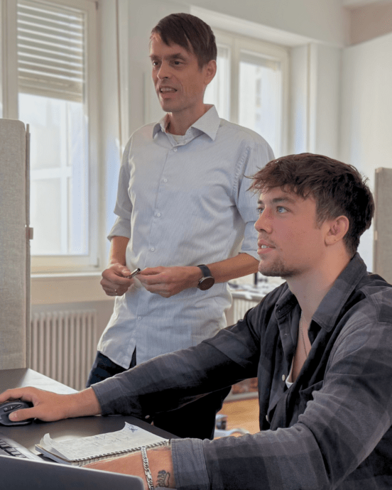 Zwei Männer arbeiten am Computer. Der sitzende Mann bedient Maus und Tastatur, der andere steht daneben und spricht.