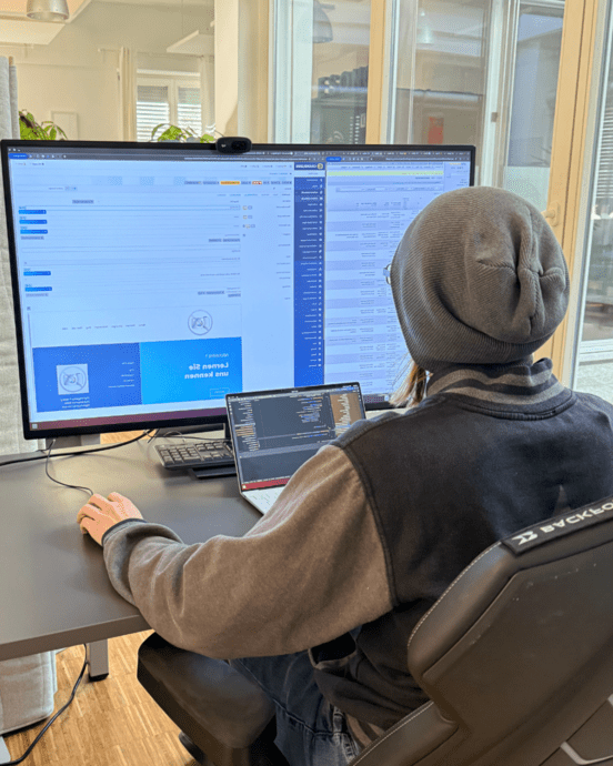 Person in Bürokleidung arbeitet an einem Schreibtisch mit zwei Bildschirmen und einem Laptop. Der Raum ist gut beleuchtet.