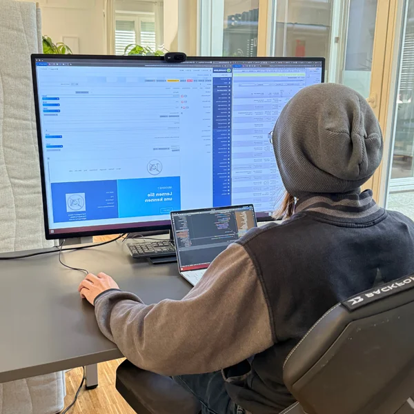 Person in Bürokleidung arbeitet an einem Schreibtisch mit zwei Bildschirmen und einem Laptop. Der Raum ist gut beleuchtet.
