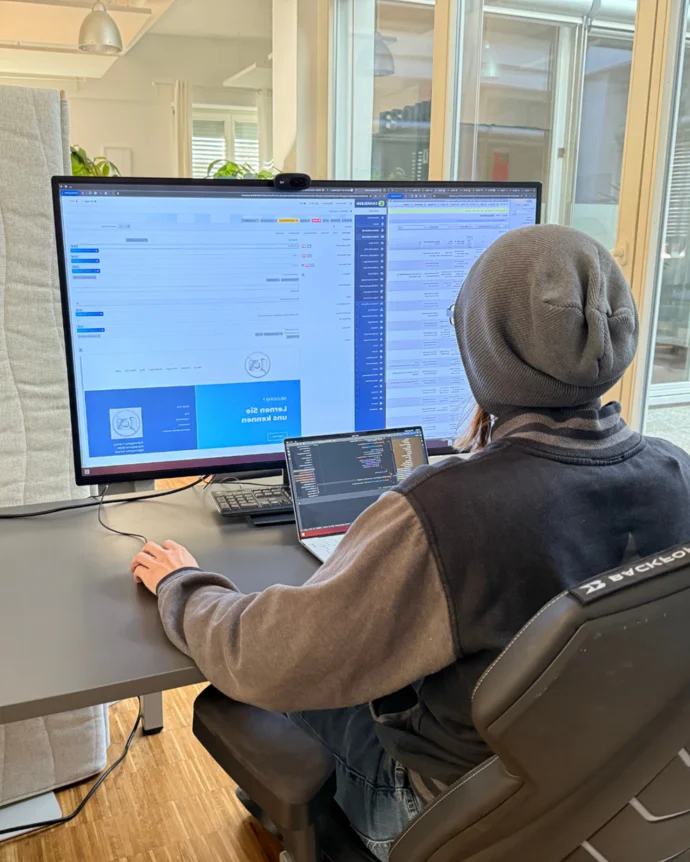 Person in Bürokleidung arbeitet an einem Schreibtisch mit zwei Bildschirmen und einem Laptop. Der Raum ist gut beleuchtet.