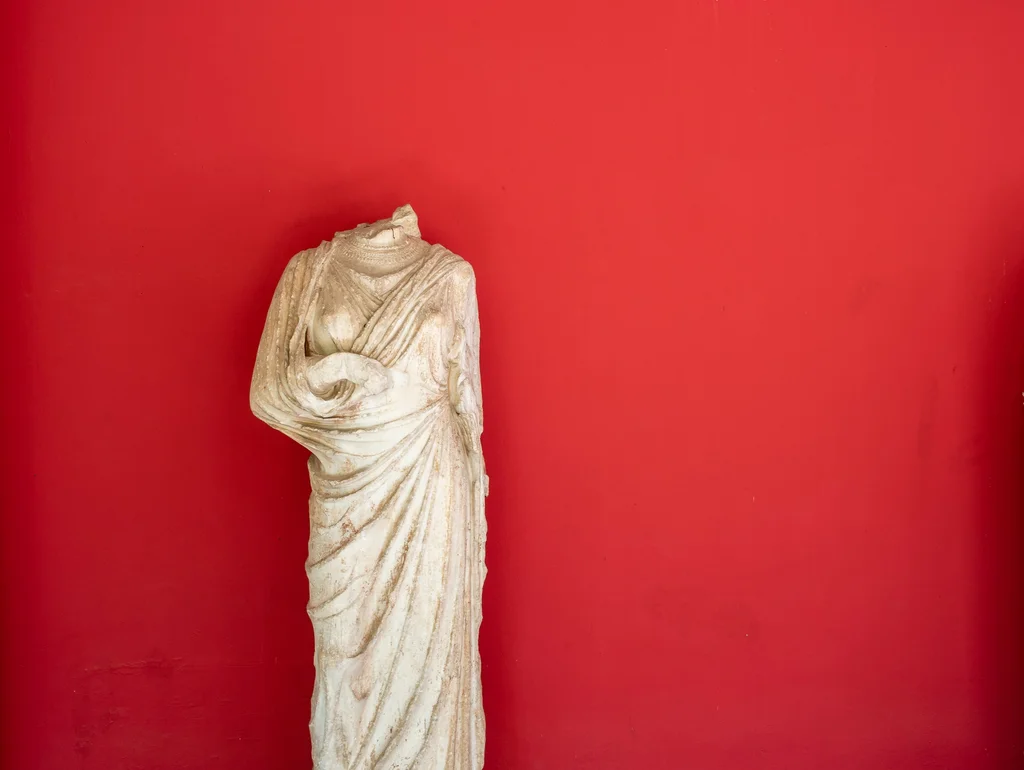 Antike kopflose Statue in drapiertem Gewand vor roter Wand.