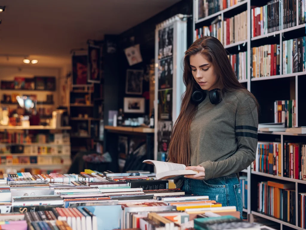 Frau liest ein Buch in einer Buchhandlung, umgeben von Bücherregalen und Tischen voller Bücher.