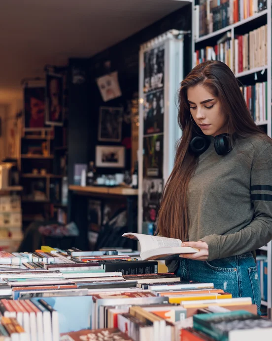 Frau liest ein Buch in einer Buchhandlung, umgeben von Bücherregalen und Tischen voller Bücher.