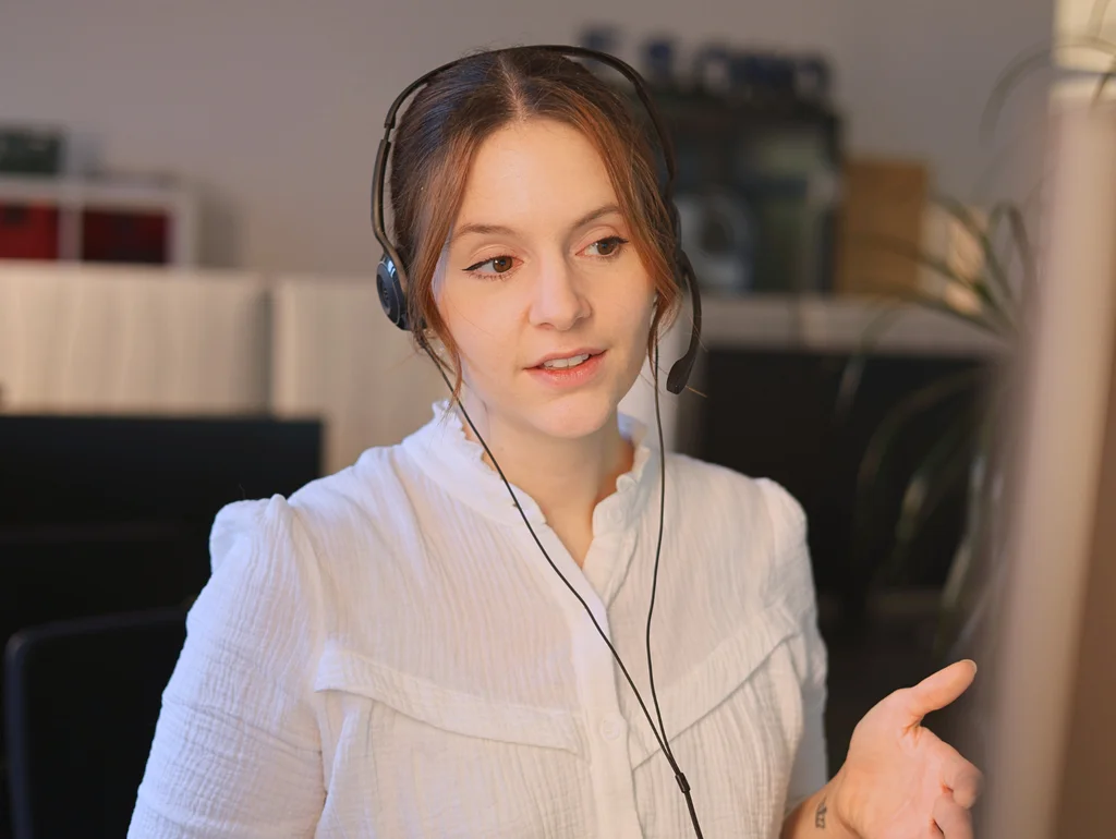Frau mit Headset arbeitet am Computer in einem Büro. Konzentrierter Ausdruck, helle Lichtverhältnisse, moderner Arbeitsplatz.