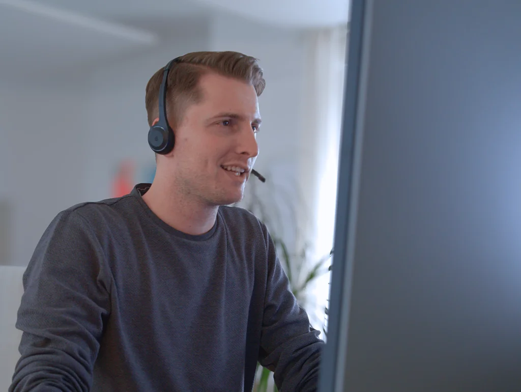 Person mit Headset arbeitet am Computer, lächelnd, in modernem Büroambiente.