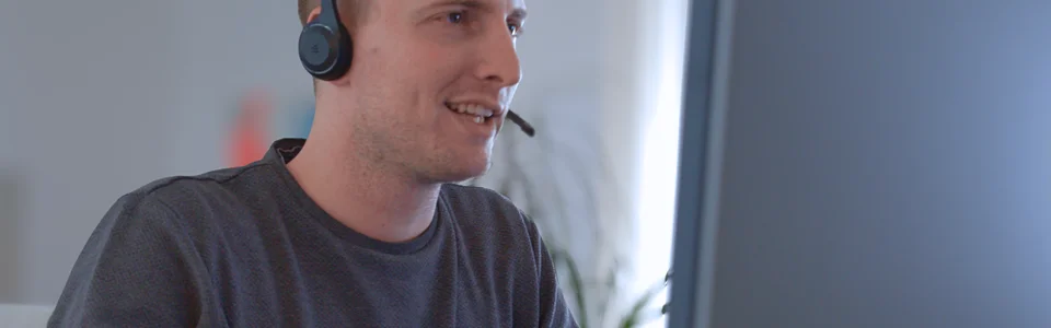 Person mit Headset arbeitet am Computer, lächelnd, in modernem Büroambiente.