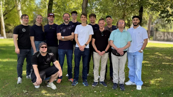 Gruppe von 13 Personen posiert im Freien vor Bäumen auf grasbewachsenem Gelände bei Tageslicht.