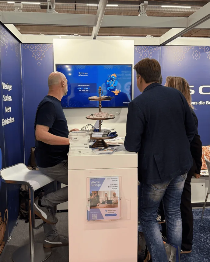 Drei Personen betrachten einen Bildschirm auf einer Messe. Ein Tisch mit Broschüren und eine Wand mit Text im Hintergrund.