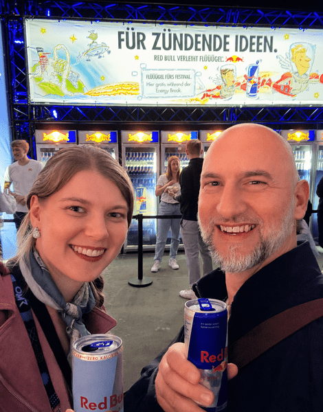 Zwei Personen lächeln in die Kamera und halten Red Bull-Dosen. Im Hintergrund ein Red Bull-Werbeschild.