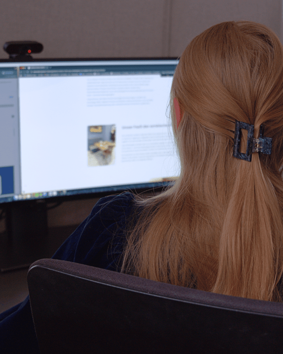 Eine Person sitzt vor zwei Monitoren mit Text und Bildern, ein Smartphone und eine Tasse stehen daneben.