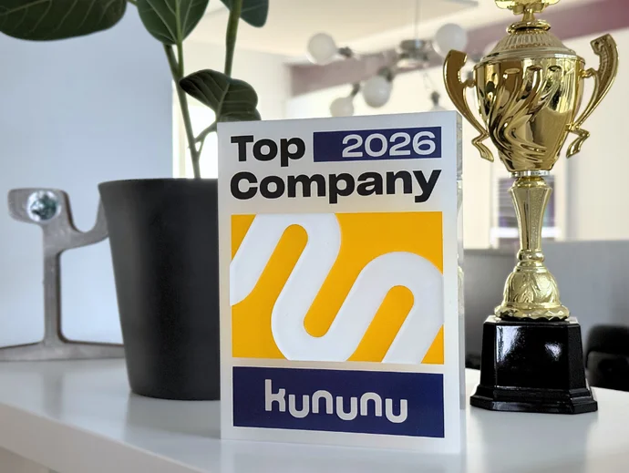 Kununu-Auszeichnung "Top Company 2026", umgeben von einer Pflanze und einem Pokal, auf einem weißen Regal.