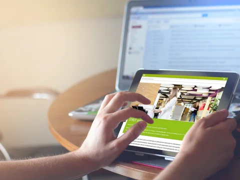 Person nutzt ein Tablet vor einem Laptop mit E-Mail-Posteingang, auf einem Tisch in heller Umgebung.