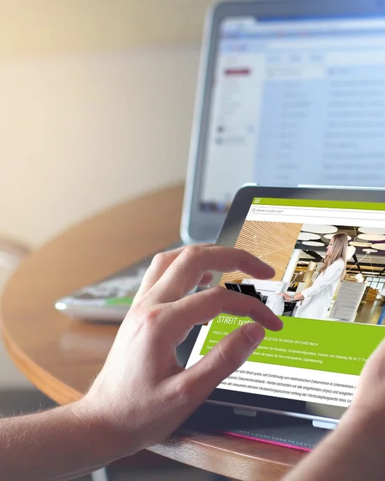 Person nutzt ein Tablet vor einem Laptop mit E-Mail-Posteingang, auf einem Tisch in heller Umgebung.