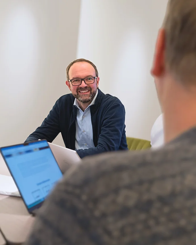 Ein lächelnder Mann in einem Meeting, Laptops auf dem Tisch, moderner Büroraum.