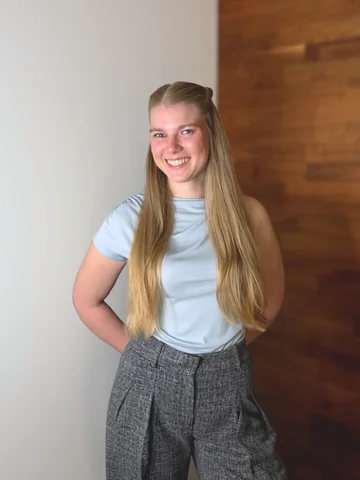 Junge Frau in grüner Bluse und blauer Jeans steht lächelnd vor holzvertäfeltem Hintergrund.