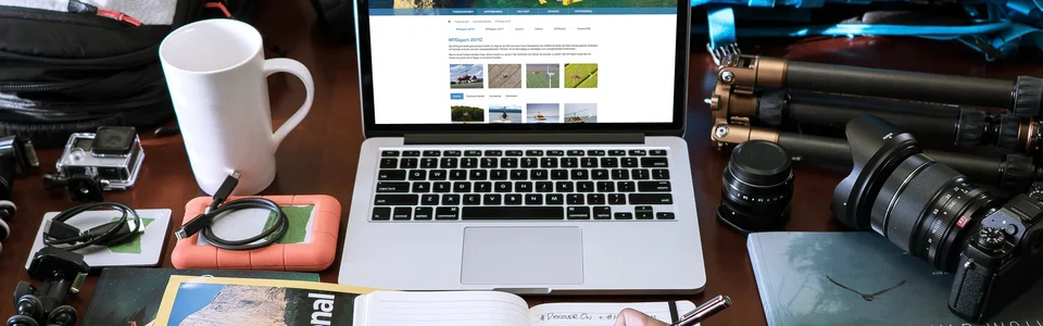 Laptop auf Schreibtisch mit Kameraausrüstung, Notizbuch, Reiseführern und Kaffeetasse, Planung für Outdoor-Fotografie.