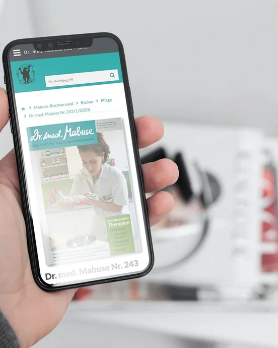 Smartphone in Hand mit aufgerufenem Magazin "Dr. med. Mabuse", im Hintergrund unscharfe Zeitschriften auf einem Tisch.