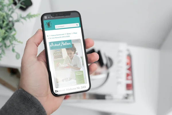 Smartphone in Hand mit aufgerufenem Magazin "Dr. med. Mabuse", im Hintergrund unscharfe Zeitschriften auf einem Tisch.