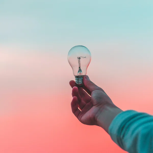 Hand hält Glühbirne vor farbigem Himmel, symbolisiert Ideen oder Innovation im kreativen Kontext.