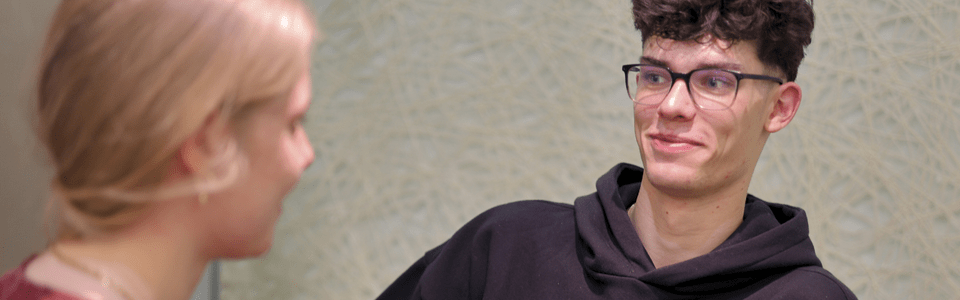 Zwei Personen im Gespräch an einem Tisch, leere Flaschen stehen vor ihnen. Eine trägt einen Hoodie, die andere hat blondes Haar.