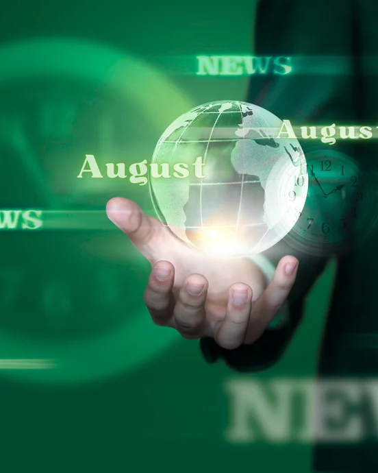 Hand hält schwebende Weltkugel mit „August“ und „News“ davor; grüne, verschwommene Uhren im Hintergrund.