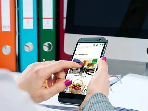 Person nutzt ein Smartphone vor einem Computerbildschirm, im Hintergrund sind bunte Ordner zu sehen.