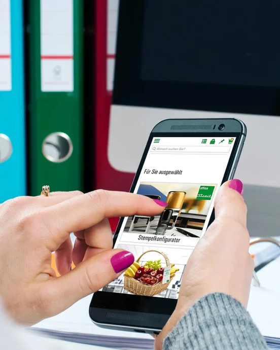 Person nutzt ein Smartphone vor einem Computerbildschirm, im Hintergrund sind bunte Ordner zu sehen.