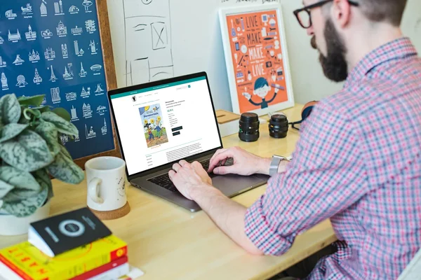 Person arbeitet am Laptop auf einem Schreibtisch mit Büchern, Pflanzen und Kameralinsen. An der Wand hängen Poster.