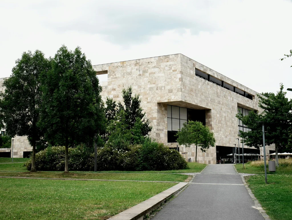 Moderne Bibliothek mit Steinfassade, umgeben von Bäumen und Grünflächen, angrenzend an einen Gehweg unter bewölktem Himmel.