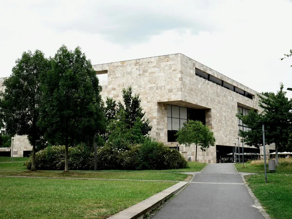 Moderne Bibliothek mit Steinfassade, umgeben von Bäumen und Grünflächen, angrenzend an einen Gehweg unter bewölktem Himmel.