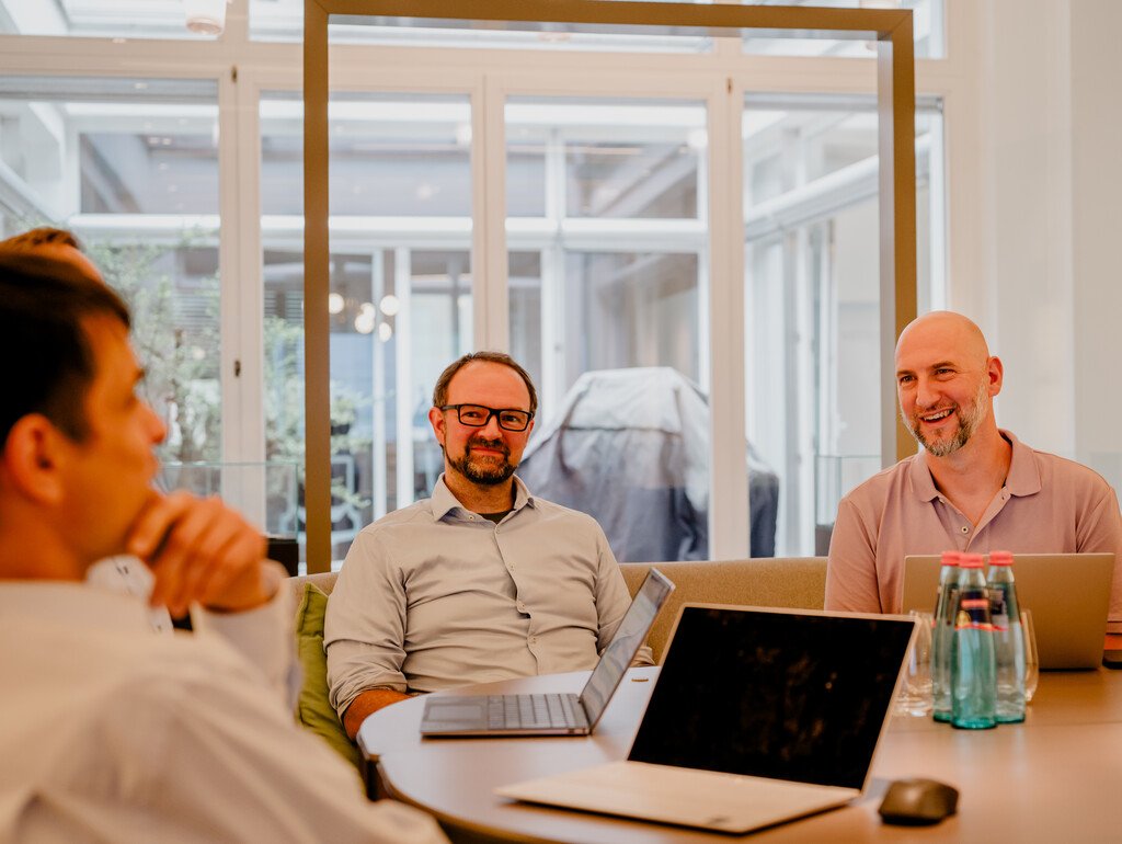 Mehrere Personen bei einem Meeting; Laptops und Wasserflaschen auf dem Tisch; moderner Konferenzraum.